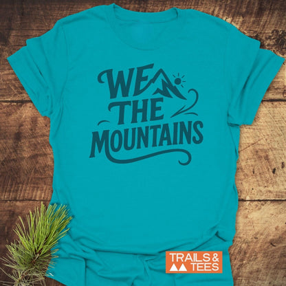 We The Mountains T-Shirt with mountain peak graphic, laid on wood. Soft, ring-spun cotton, pre-shrunk, durable print. Includes “TRAILS & TEES” logo and pine sprig.