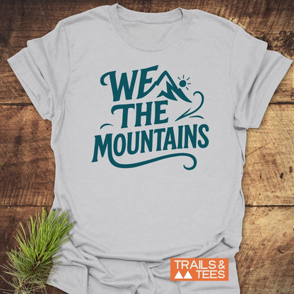 We The Mountains T-Shirt in light-gray, features teal graphic and orange logo, laid on wood, styled with pine sprig for outdoor-themed appeal.