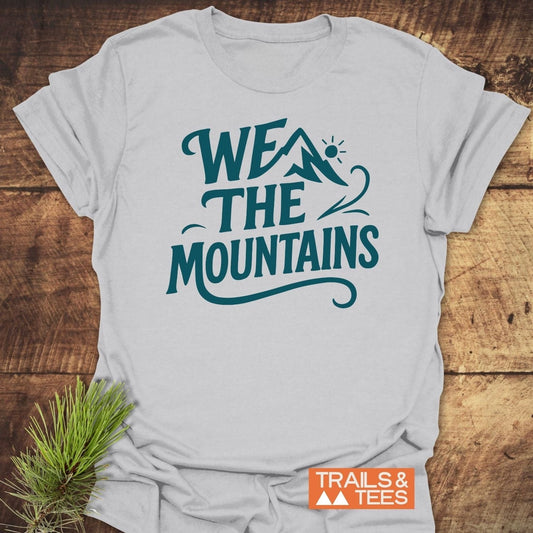 We The Mountains T-Shirt in light-gray, features teal graphic and orange logo, laid on wood, styled with pine sprig for outdoor-themed appeal.