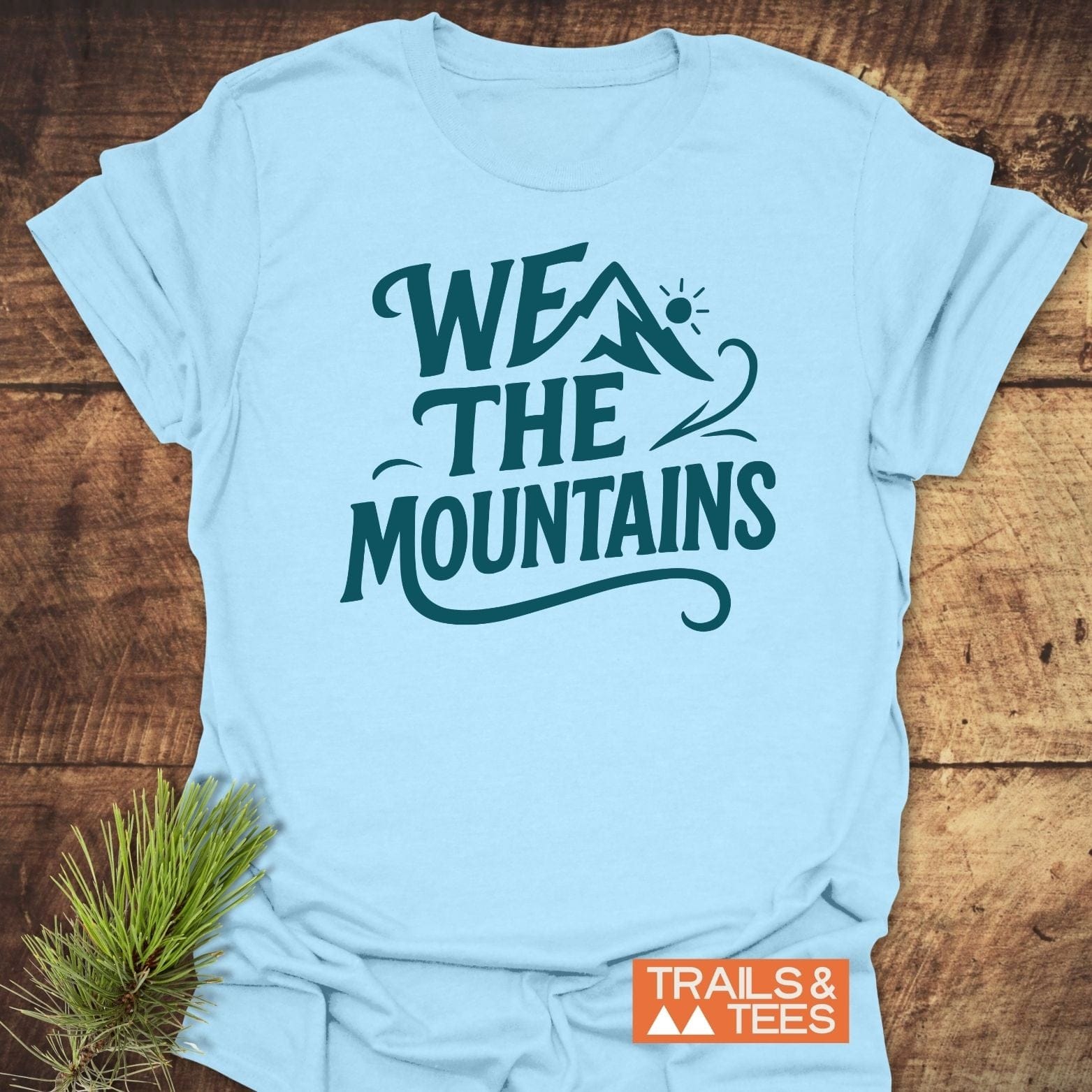 We The Mountains T-Shirt featuring a stylized mountain and sun graphic, crafted from soft ring-spun cotton, displayed flat on a wooden surface.
