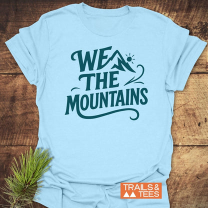 We The Mountains T-Shirt featuring a stylized mountain and sun graphic, crafted from soft ring-spun cotton, displayed flat on a wooden surface.