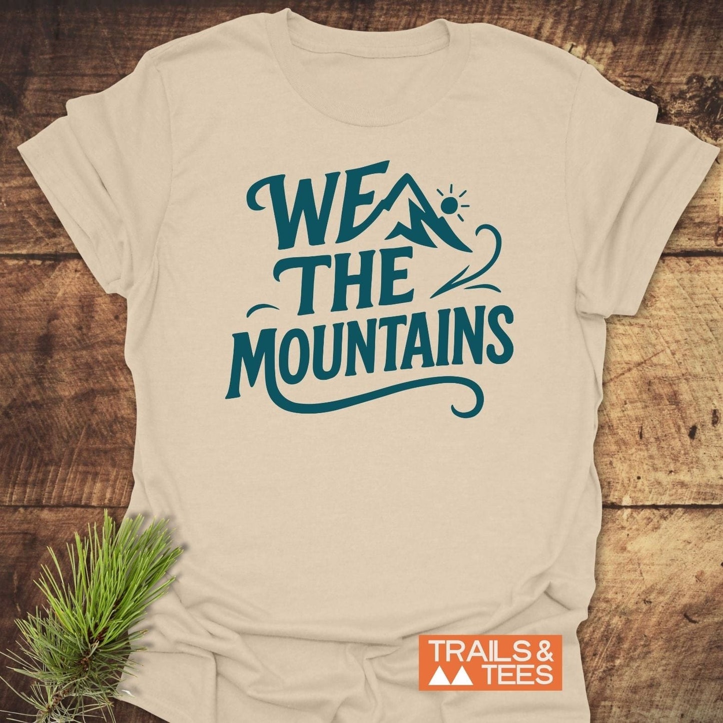 We The Mountains T-Shirt on wooden surface, featuring teal slogan and mountain icon. Lightweight, ring-spun cotton, durable print. Includes sprig of pine and Trails & Tees logo.