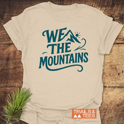 We The Mountains T-Shirt on wooden surface, featuring teal slogan and mountain icon. Lightweight, ring-spun cotton, durable print. Includes sprig of pine and Trails & Tees logo.