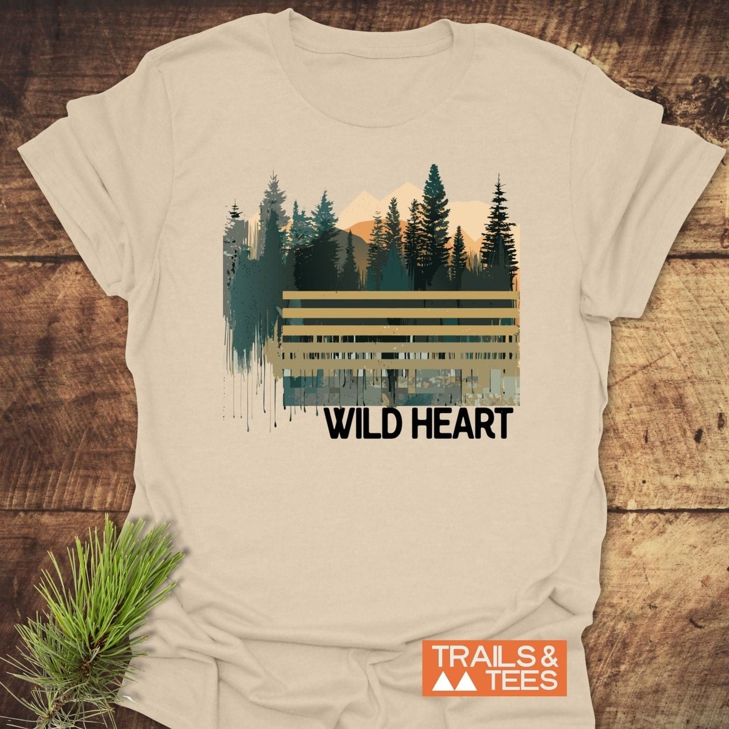 Wild Heart Graphic T-Shirt featuring a mountain-and-forest design, displayed on wood with pine sprig. Made from soft, durable cotton for outdoor enthusiasts.