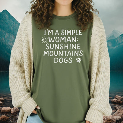 Woman wearing Sunshine Mountains Dogs T-Shirt with olive-green color, casual font. Paired with light jeans and cream cardigan, set against a mountain-lake backdrop.
