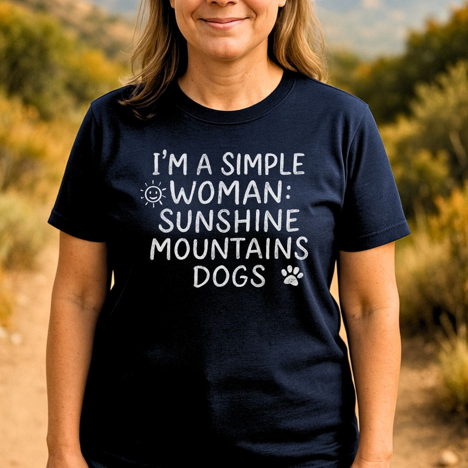 Person wearing a navy Sunshine Mountains Dogs T-shirt with white chalk-style text and icons, standing outdoors on a dirt path with hills in the background.
