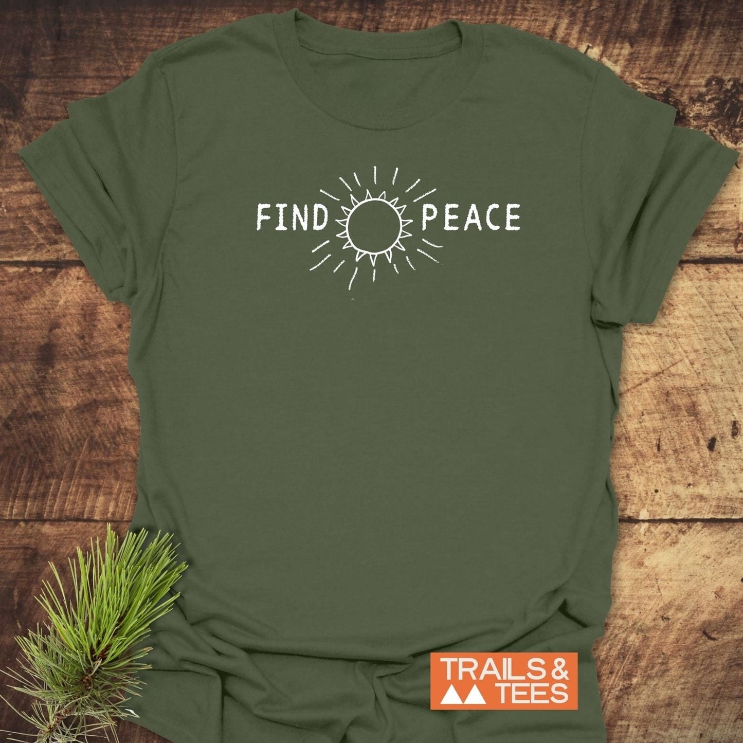 Forest-green Find Peace T-Shirt with white sun graphic on chest, laying flat with TRAILS & TEES badge, and pine sprig beside it.