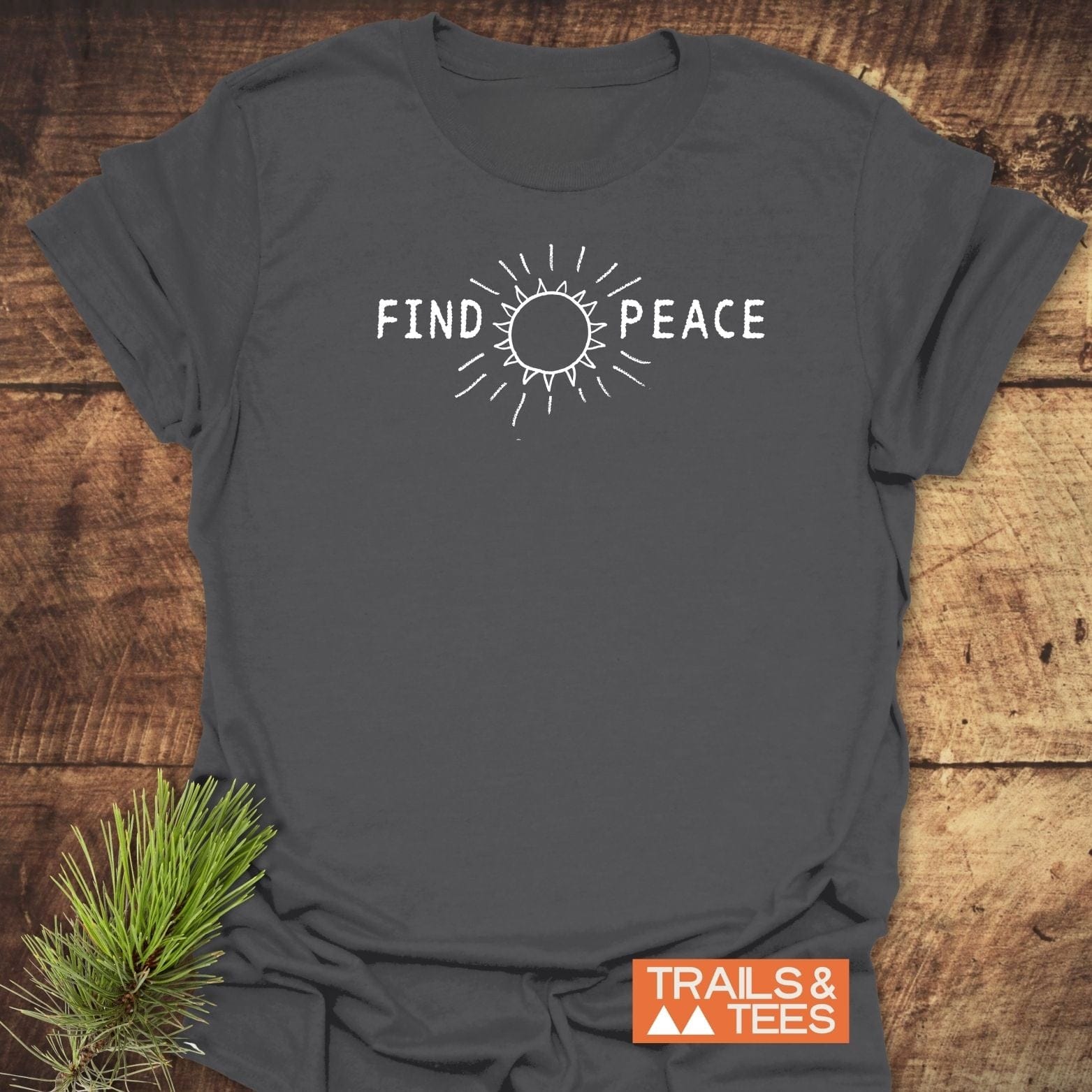 Find Peace T-Shirt featuring a sun line-drawing and FIND PEACE text, laid on wood with pine sprig, showcasing Trails & Tees logo.
