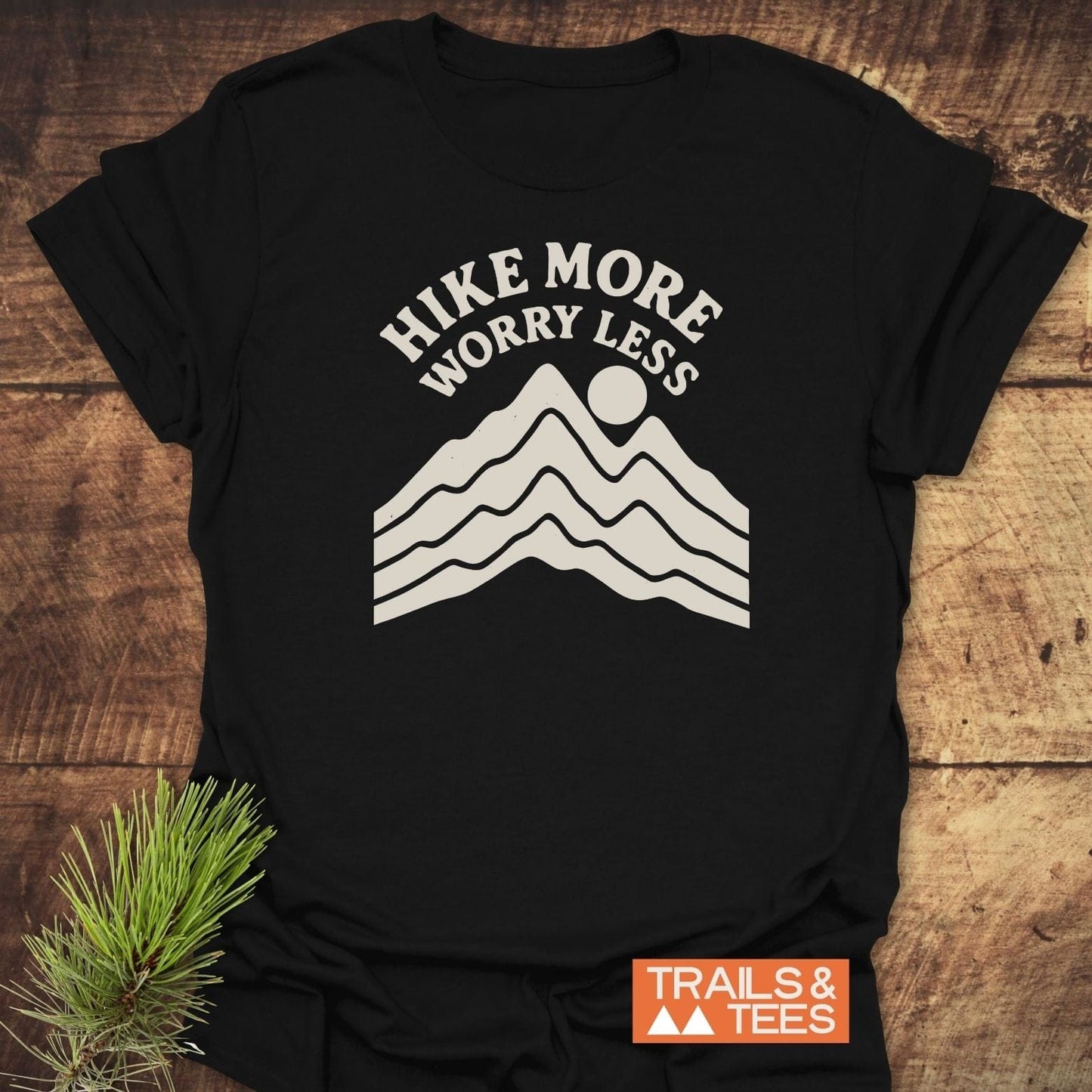 Hike More Worry Less T-Shirt with mountain and sun graphic, laid flat on wood, pine sprig nearby, featuring durable print and soft, ring-spun cotton.