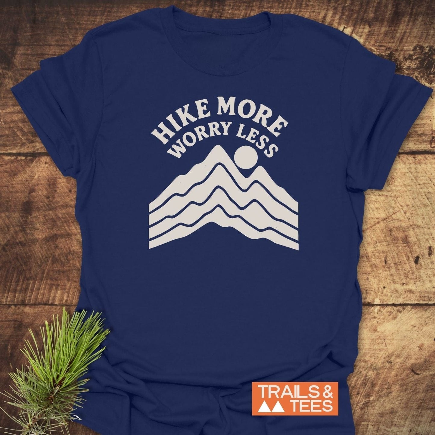 Navy-blue Hike More Worry Less T-shirt with a white mountain graphic, designed by Trails & Tees, laid on rustic wood with a pine sprig.