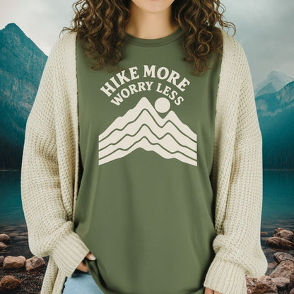Person in Hike More Worry Less T-Shirt with mountain graphic, wearing a cardigan, standing in front of a serene mountain-lake backdrop.