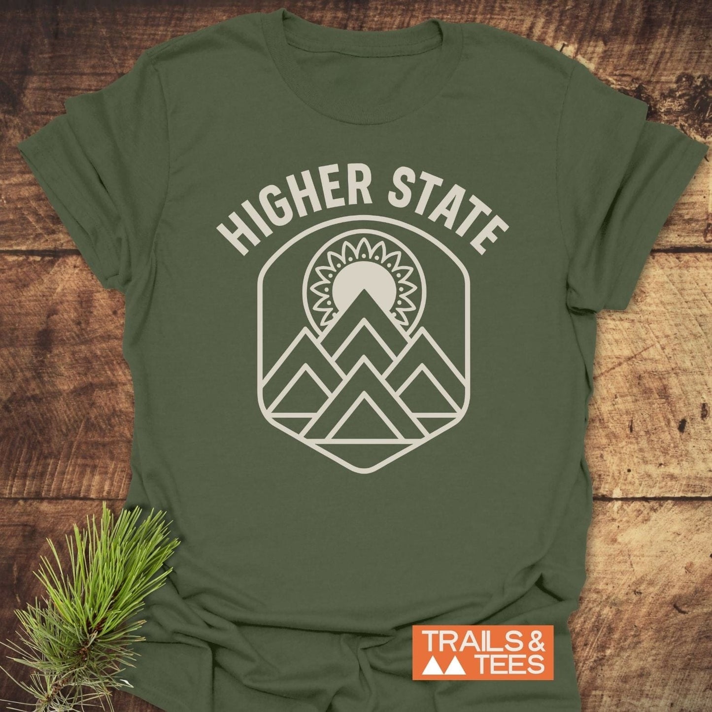 Higher State Mountain Mandala T-Shirt on wood, featuring a mountain and sun graphic. Includes TRAILS & TEES label, highlighting adventure-inspired design and durable print.