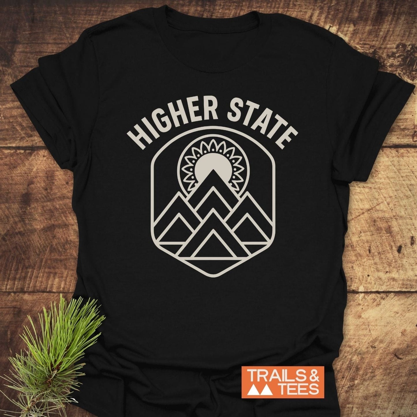 Higher State Mountain Mandala T-Shirt on wood, featuring mountain and sunburst badge. Orange TRAILS & TEES box and pine branch accentuate outdoor theme.