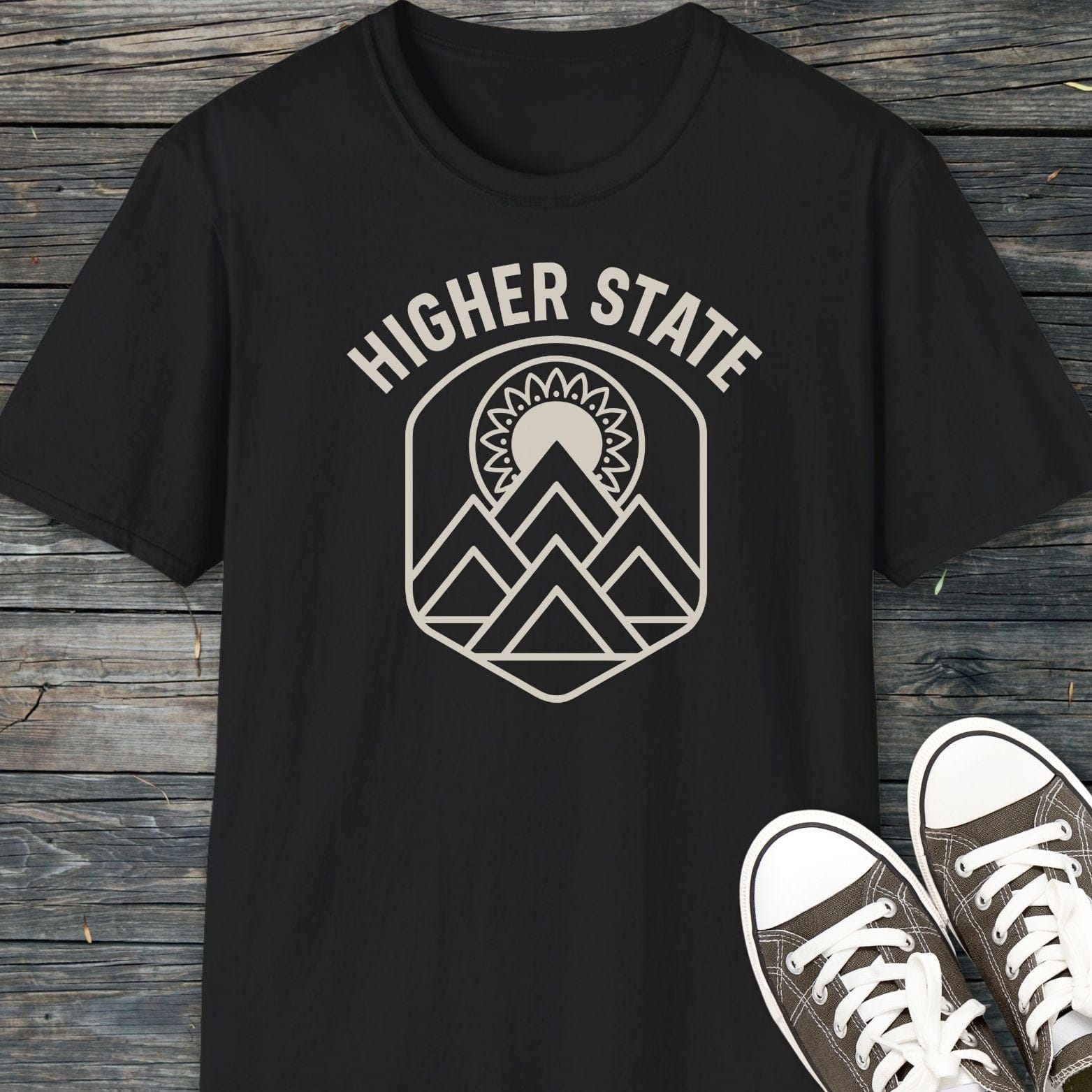 Higher State Mountain Mandala T-Shirt, flat-lay on wood, with a mountain-and-sun emblem; includes black/gray canvas sneakers in view.