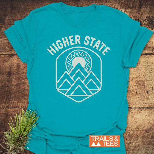 Higher State Mountain Mandala T-Shirt with a white mountain and sun graphic, laid on wood, showcasing outdoor-inspired design with TRAILS & TEES logo.