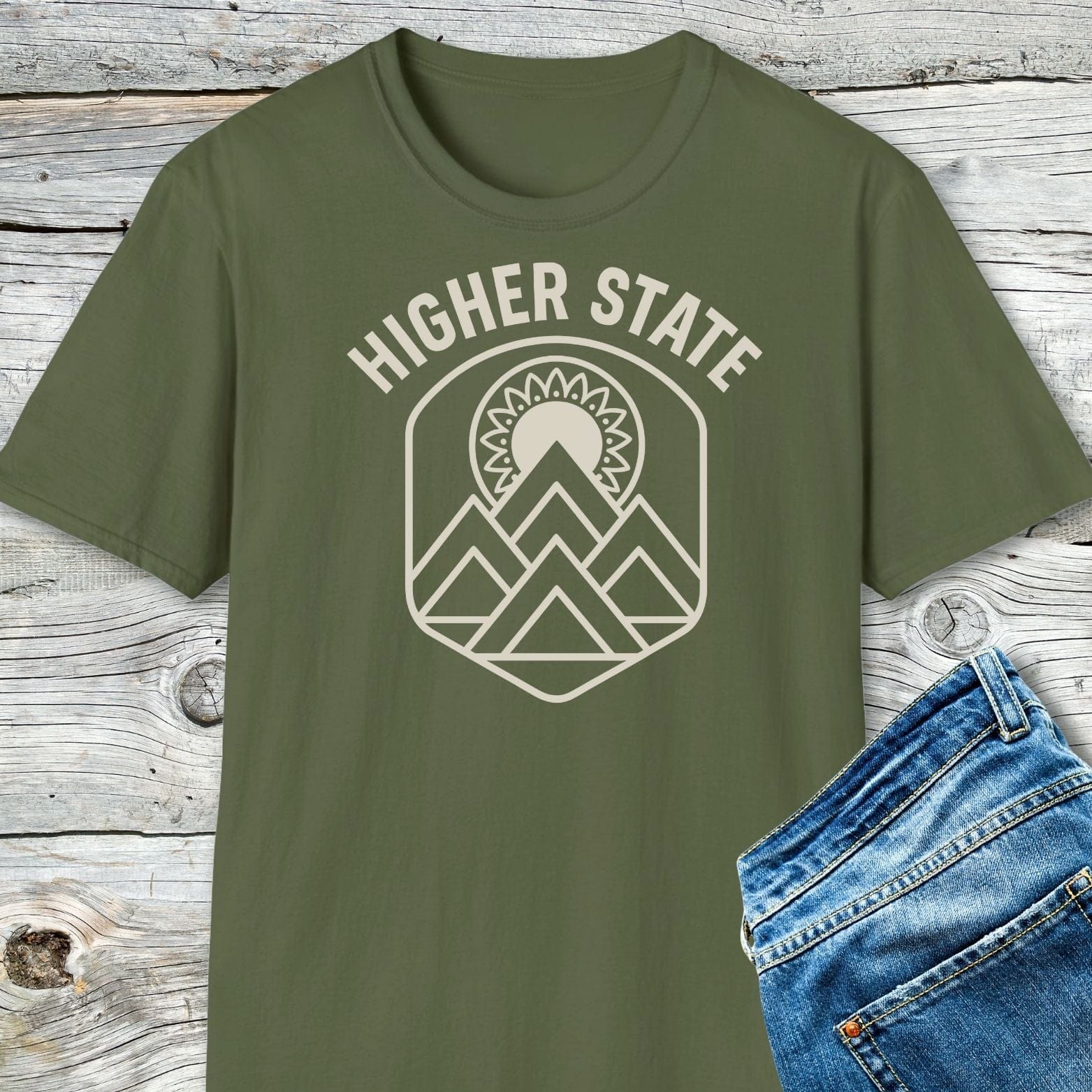 Higher State Mountain Mandala T-Shirt, olive-green, laid on wooden surface, featuring cream mountain-and-sun graphic. Partially visible blue denim jeans to the right.