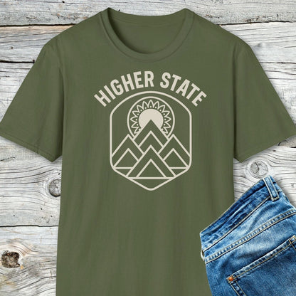 Higher State Mountain Mandala T-Shirt, olive-green, laid on wooden surface, featuring cream mountain-and-sun graphic. Partially visible blue denim jeans to the right.