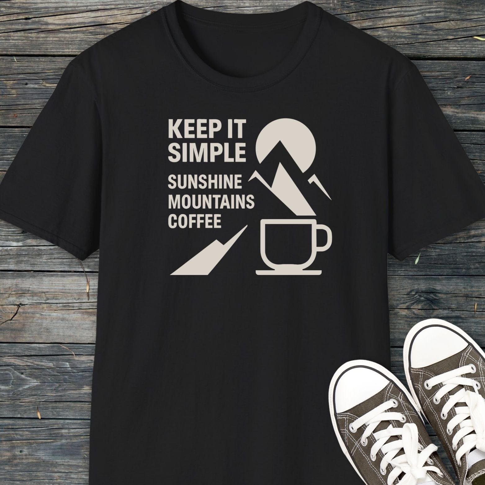 Keep It Simple Mountain T-Shirt with “KEEP IT SIMPLE” graphic, sun-mountain-coffee icon, and classic low-top dark canvas sneakers on a wooden surface.