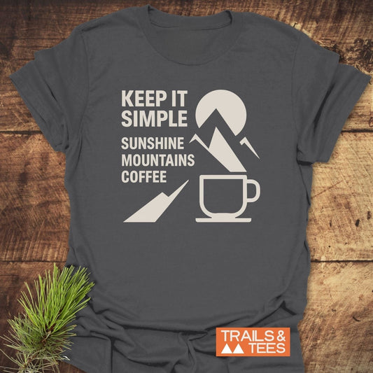 Keep It Simple Mountain T-Shirt featuring a white graphic of sun, mountains, and coffee cup, with TRAILS & TEES logo, laid on wood.