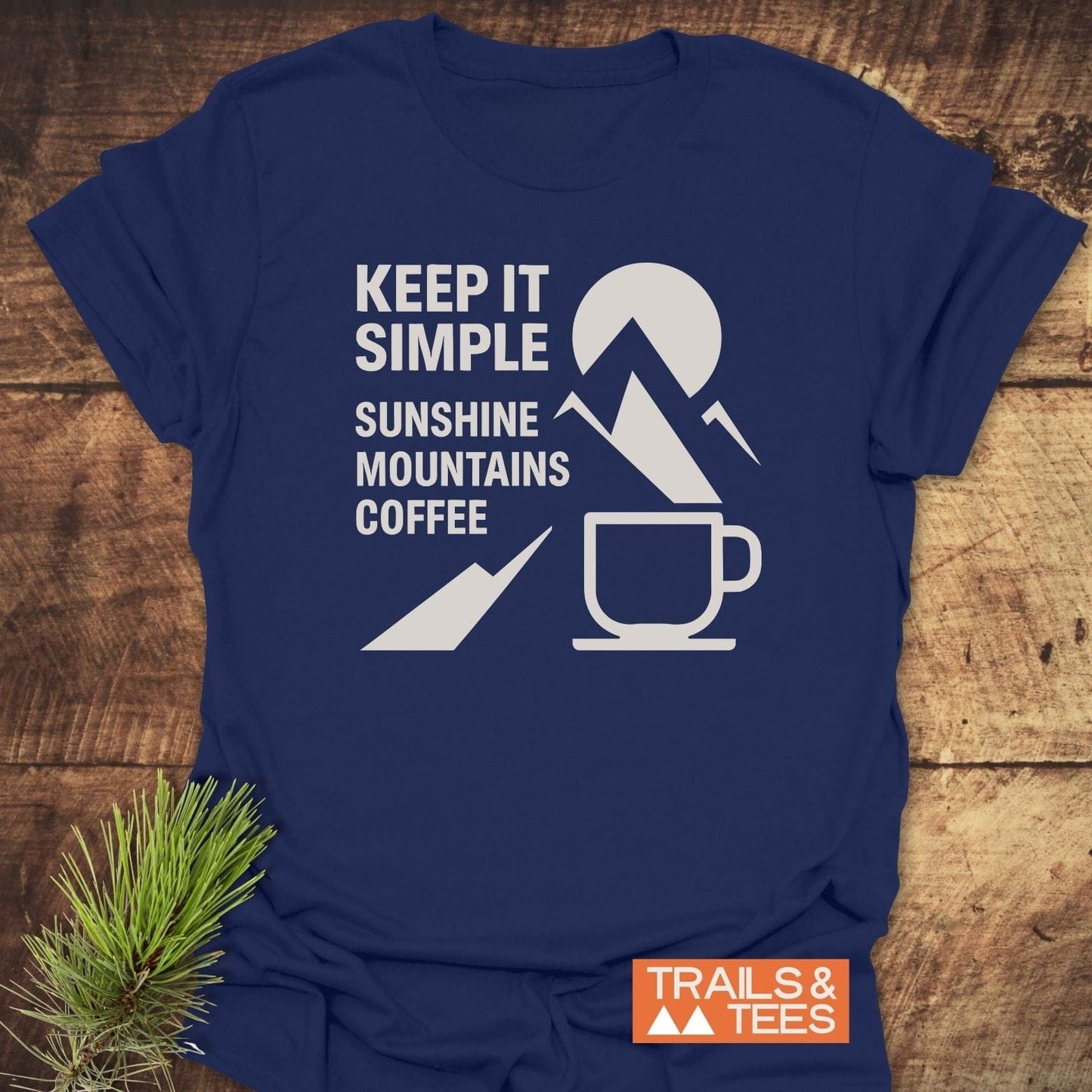 Keep It Simple Mountain T-Shirt with SUNSHINE • MOUNTAINS • COFFEE print, featuring sun and mountains graphic, laid on wood with pine sprig.