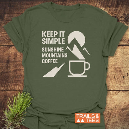 Keep It Simple Mountain T-Shirt with “KEEP IT SIMPLE” and icons, showcasing a nature theme, laid on a wooden surface with pine needles beside it.