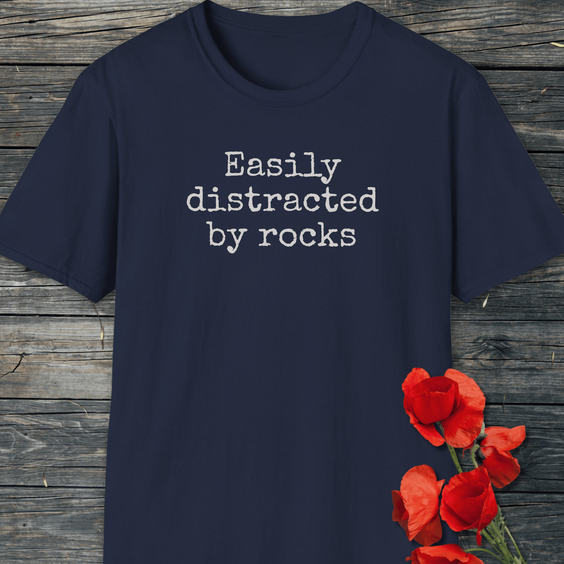 Easily Distracted By Rocks T-Shirt on wooden surface, featuring white typewriter-style text, with red poppy flowers nearby. Soft, durable, ring-spun cotton for outdoor adventures.