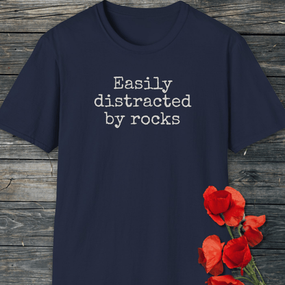 Easily Distracted By Rocks T-Shirt on wooden surface, featuring white typewriter-style text, with red poppy flowers nearby. Soft, durable, ring-spun cotton for outdoor adventures.