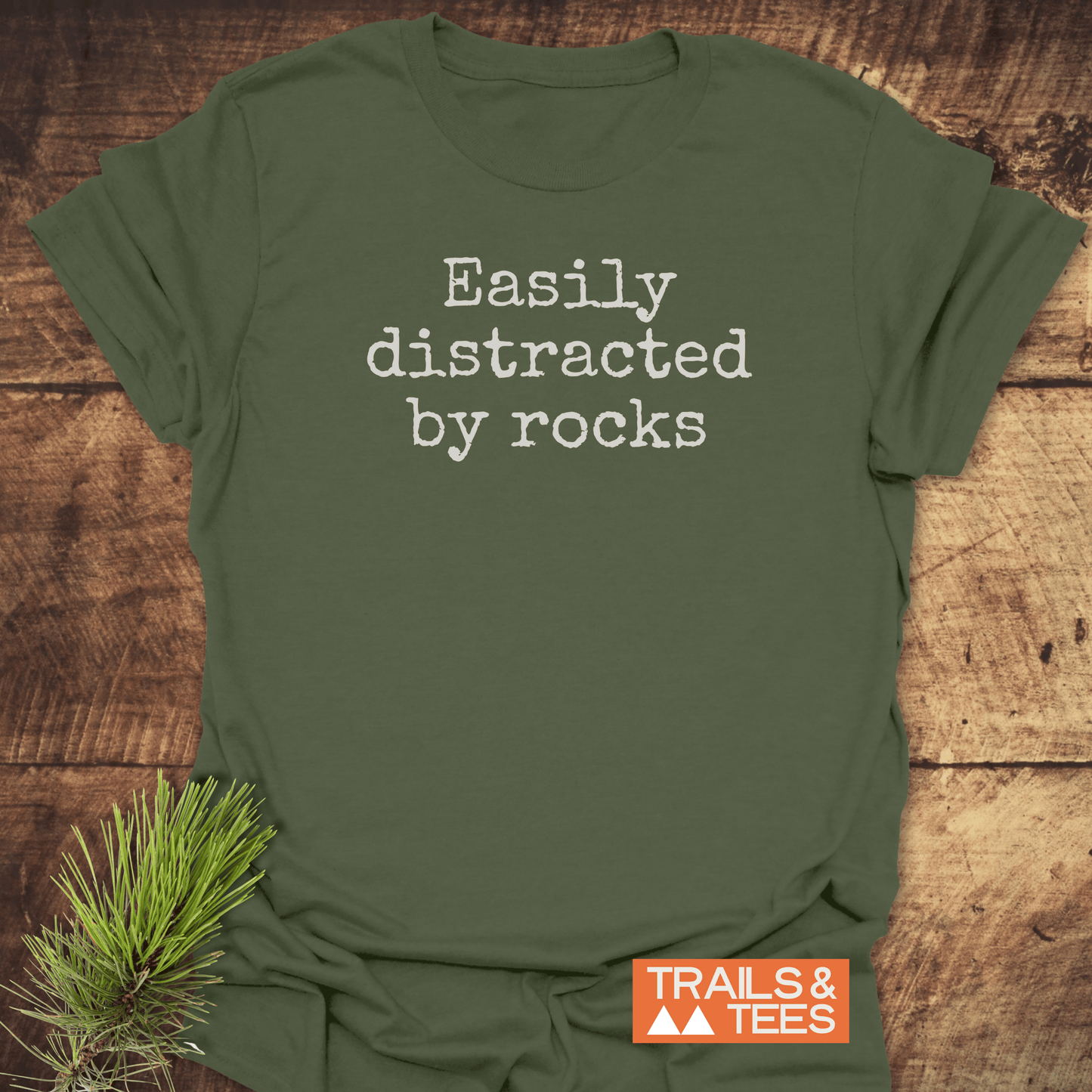 Easily Distracted By Rocks T-Shirt with typewriter font, showcasing trail-tested comfort and durable print, laid on wood with Trails & Tees logo.