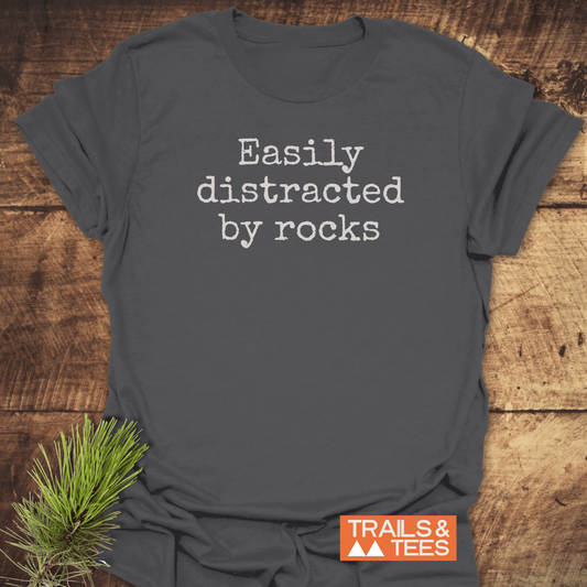 Dark gray T-shirt with “Easily Distracted By Rocks” text in typewriter font, featuring a small orange-and-white “TRAILS & TEES” logo on rustic wood.