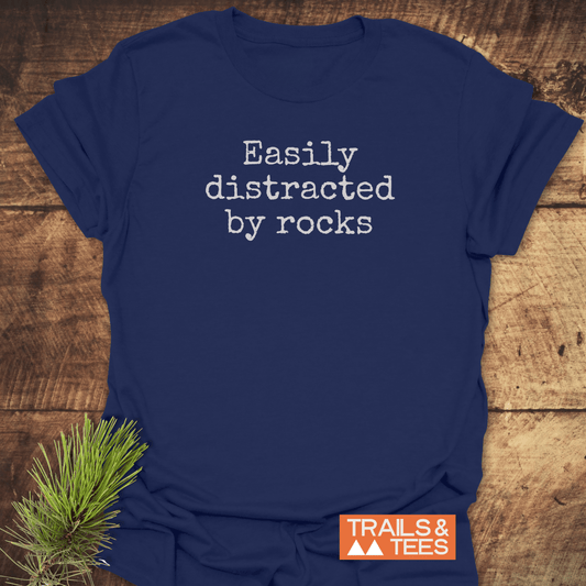 Navy blue T-shirt with Easily distracted by rocks in white font, featuring a small pine branch accent and Trails & Tees logo.