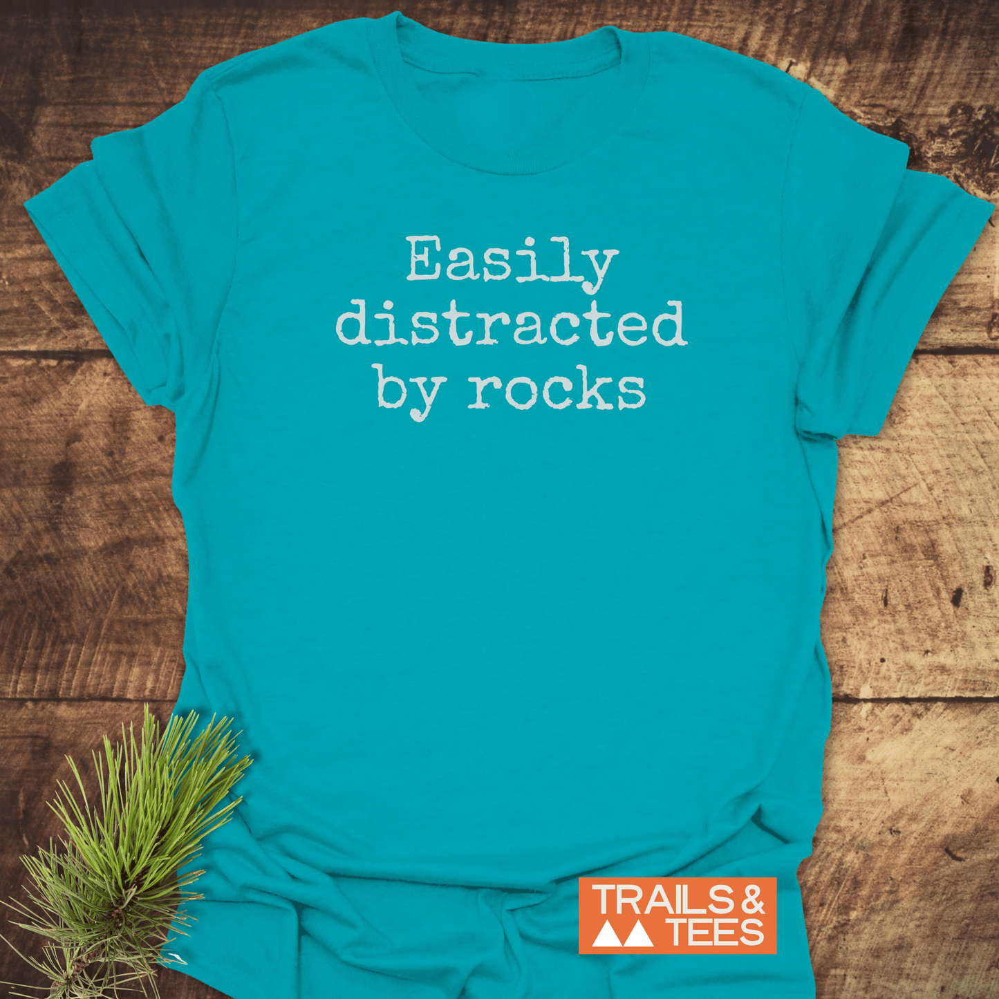 Teal T-shirt reads “Easily distracted by rocks” in white typewriter font, featuring TRAILS & TEES logo. Made of soft, durable ring-spun cotton.
