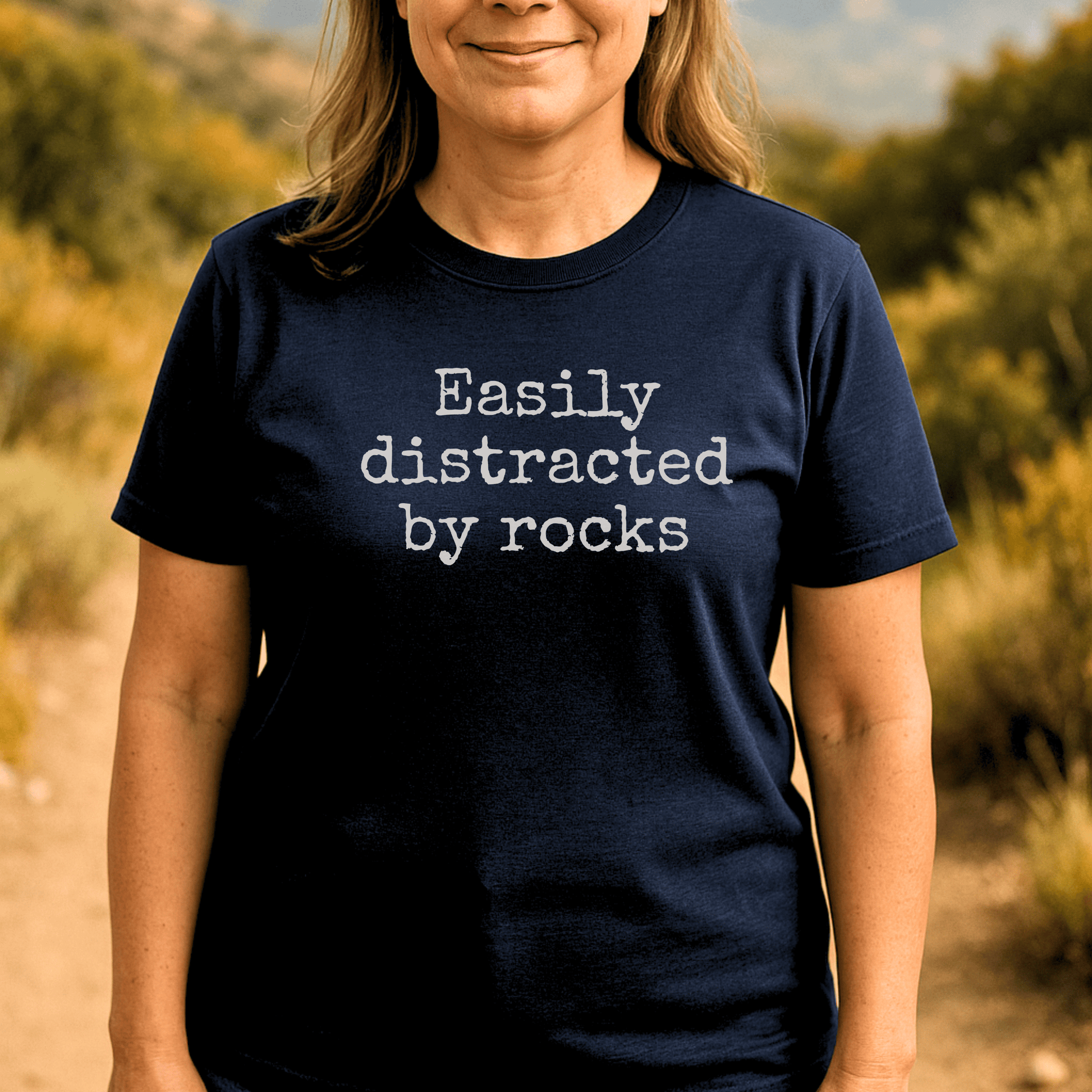 Person outdoors wearing the Easily Distracted By Rocks T-Shirt, navy blue with white text, showcasing comfort and durability for outdoor enthusiasts.