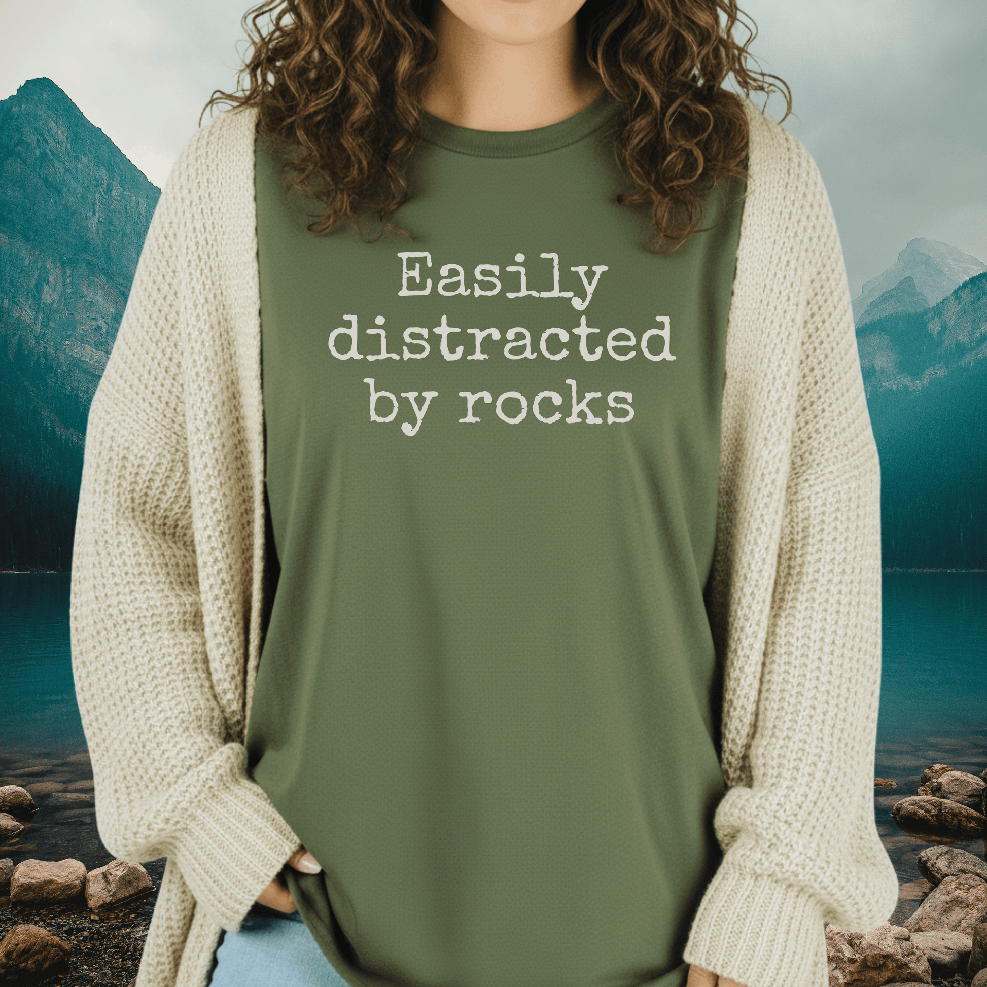 Person wearing Easily Distracted By Rocks T-shirt under a chunky knit cardigan, near a mountain lake, embodying outdoor adventure and comfort.