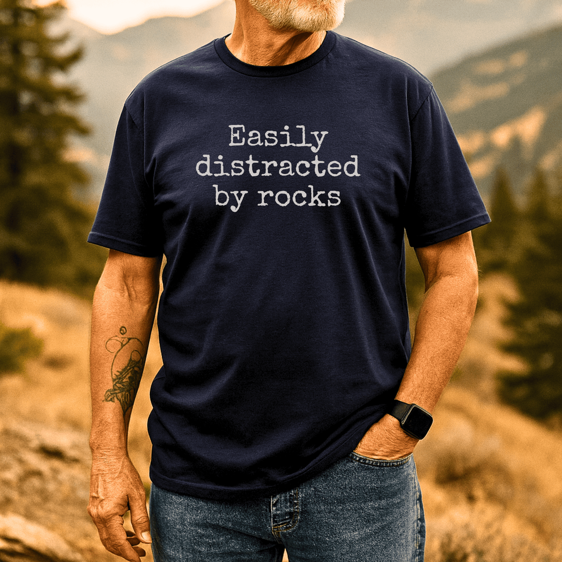Man outdoors wearing the Easily Distracted By Rocks T-Shirt, showcasing the navy design with durable print, emphasizing adventure-ready comfort and style.