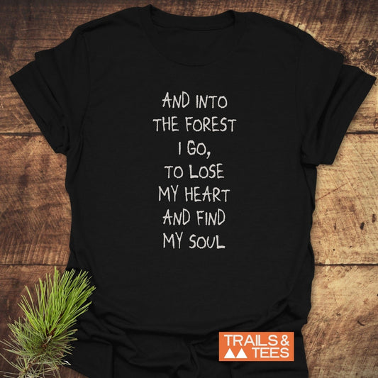 Black And Into The Forest I Go T-shirt on wood, featuring white hand-lettered text. Includes a small pine branch. Comfortable, durable, and eco-friendly design by TRAILS & TEES.