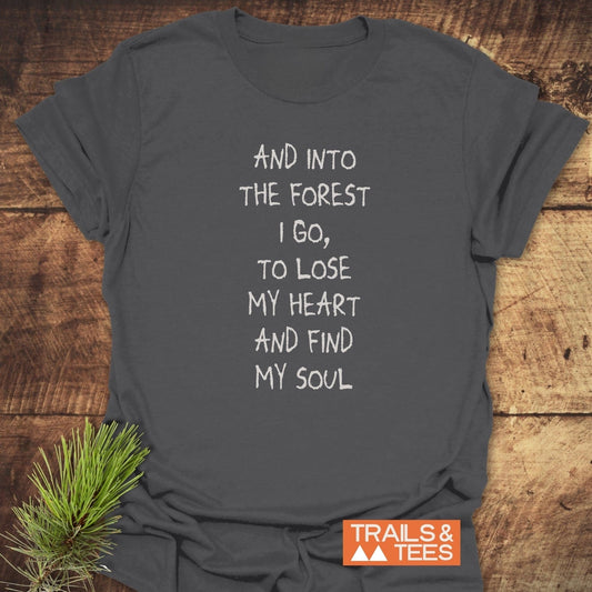 Charcoal-gray And Into The Forest I Go T-shirt with white hand-lettered design, laid on wood with pine sprig and Trails & Tees logo.