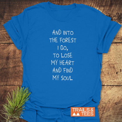 And Into The Forest I Go T-Shirt, featuring hand-drawn text, laid on wood with pine sprig. Soft, durable cotton, perfect for outdoor enthusiasts.