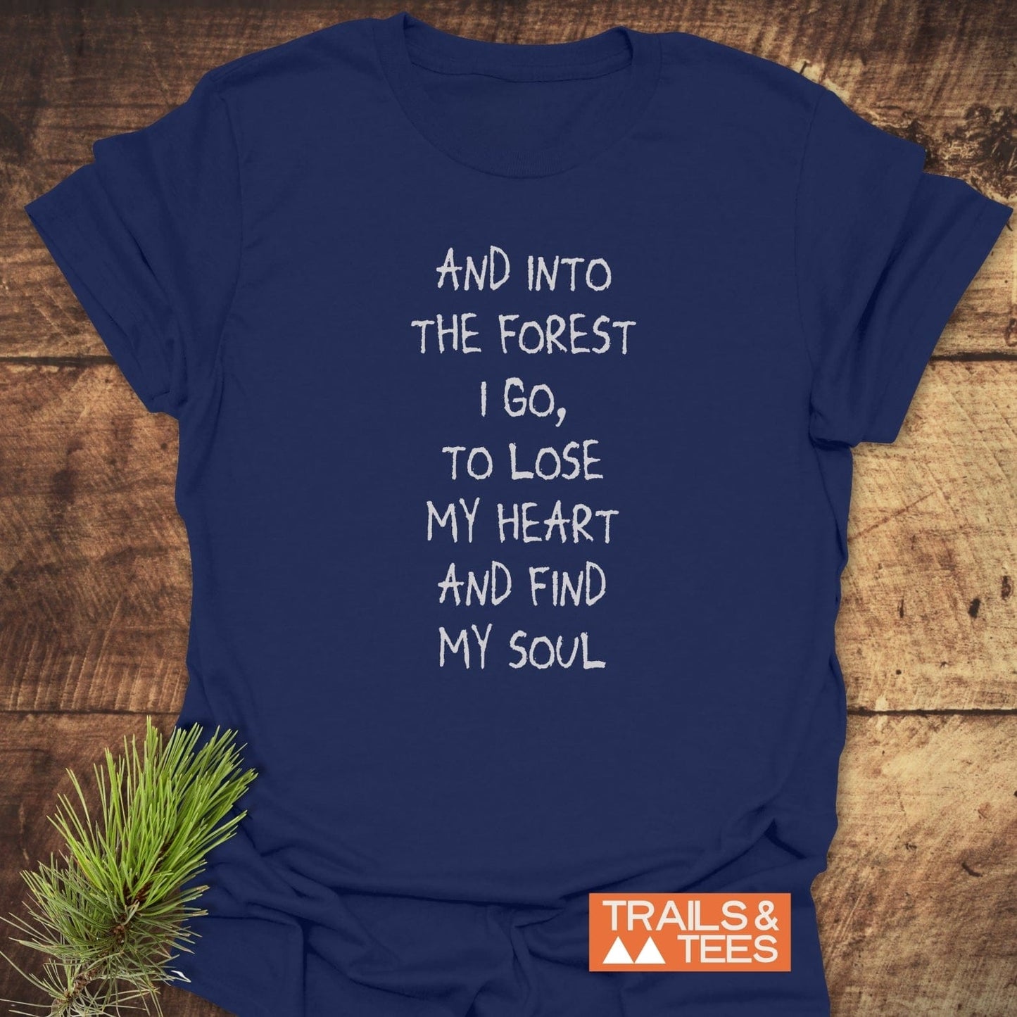Navy-blue T-shirt with quote “And into the forest I go…” in white, by TRAILS & TEES. Laid flat on wood, pine sprig accent.