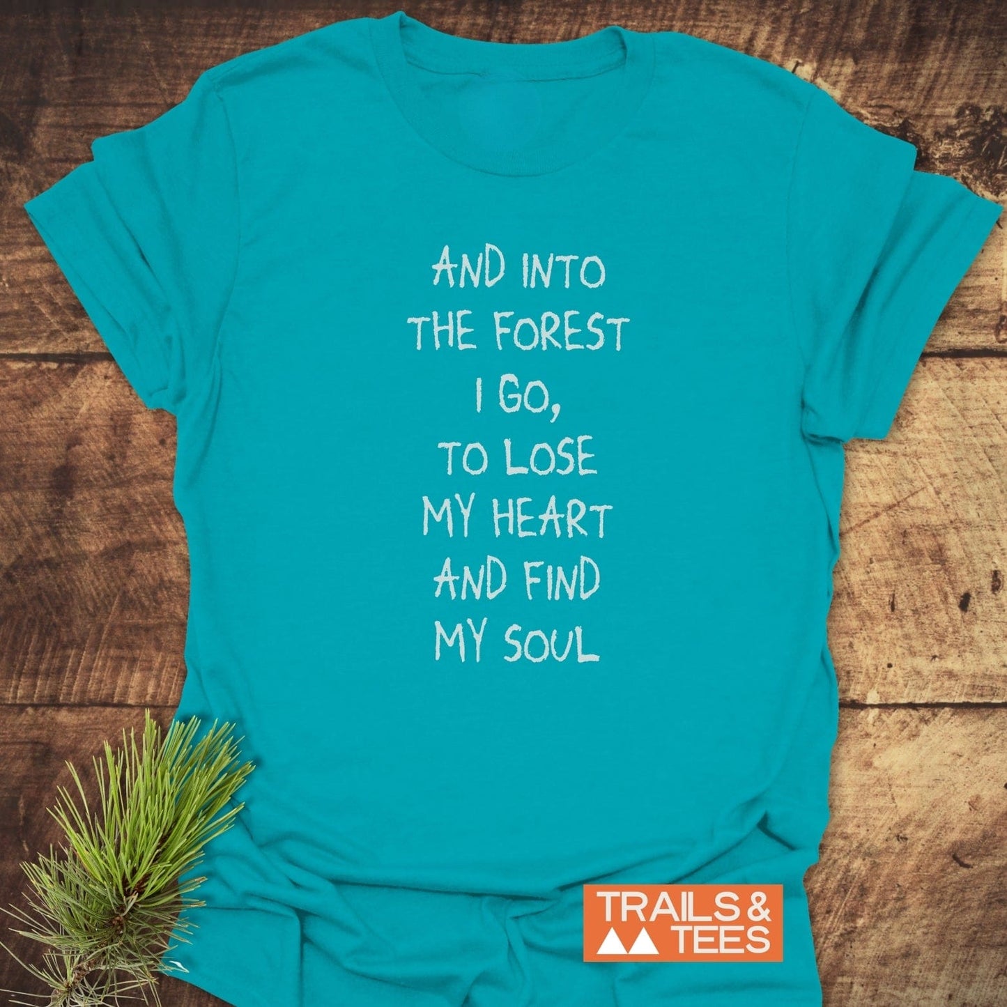 And Into The Forest I Go T-Shirt on rustic wood, featuring white hand-lettered text with “TRAILS & TEES” logo. Includes spruce twig accent.