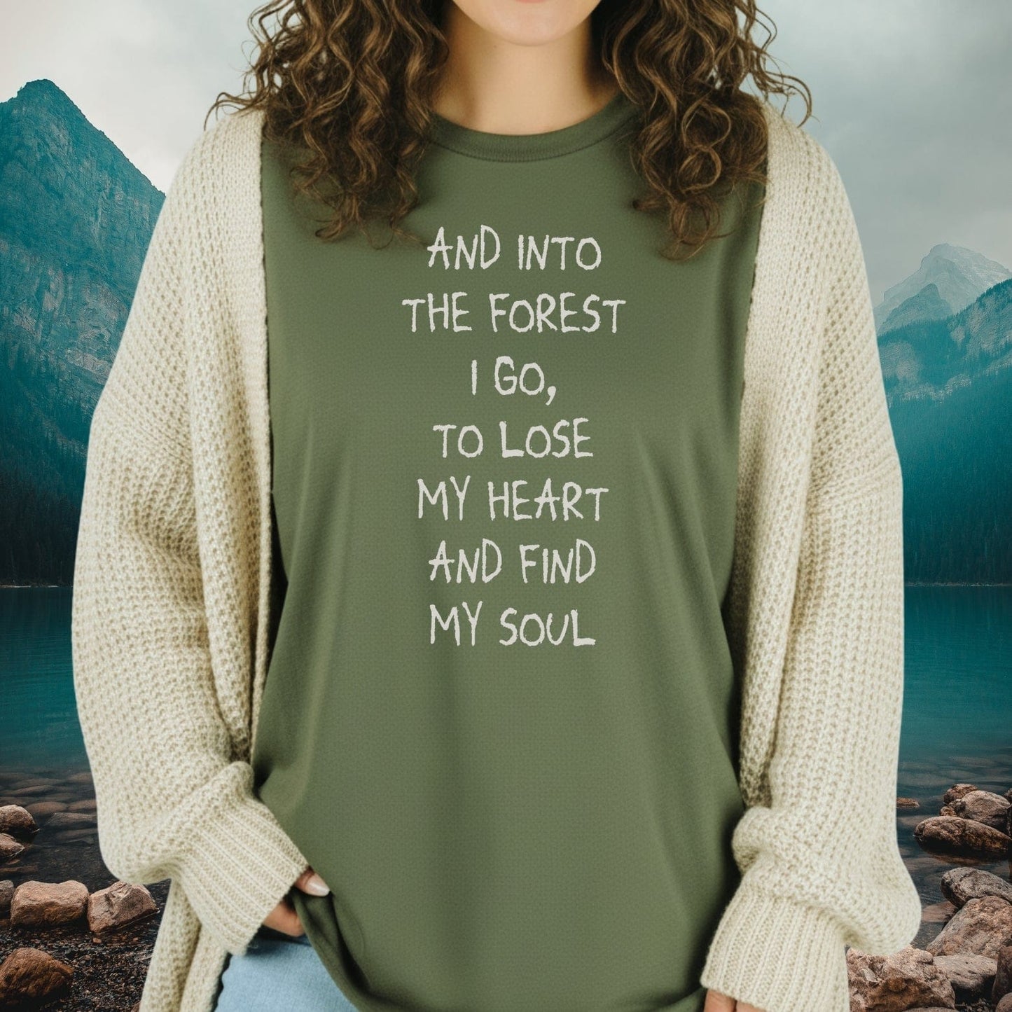 Person wearing the And Into The Forest I Go T-Shirt with a cream-knit cardigan, set against a serene mountain-lake backdrop, highlighting adventure-ready comfort and design.
