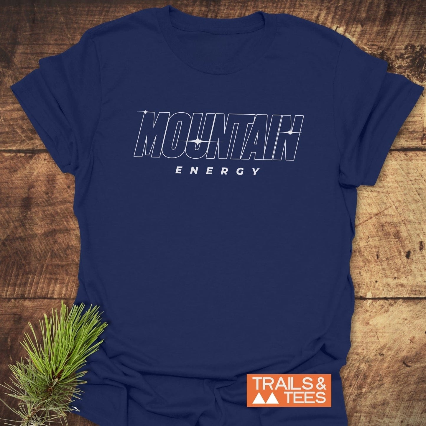 Mountain Energy T-Shirt flat-lay with bold print, placed on wood surface, pine sprig accent, featuring durable, soft cotton for outdoor enthusiasts.