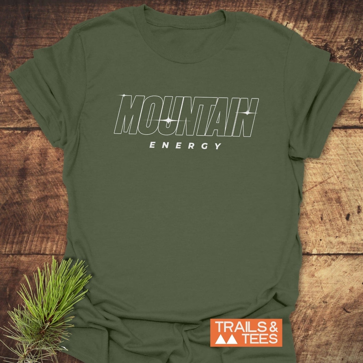 Mountain Energy T-Shirt on wooden surface, featuring outlined MOUNTAIN and solid ENERGY text, with Trails & Tees logo, and pine needle accent.