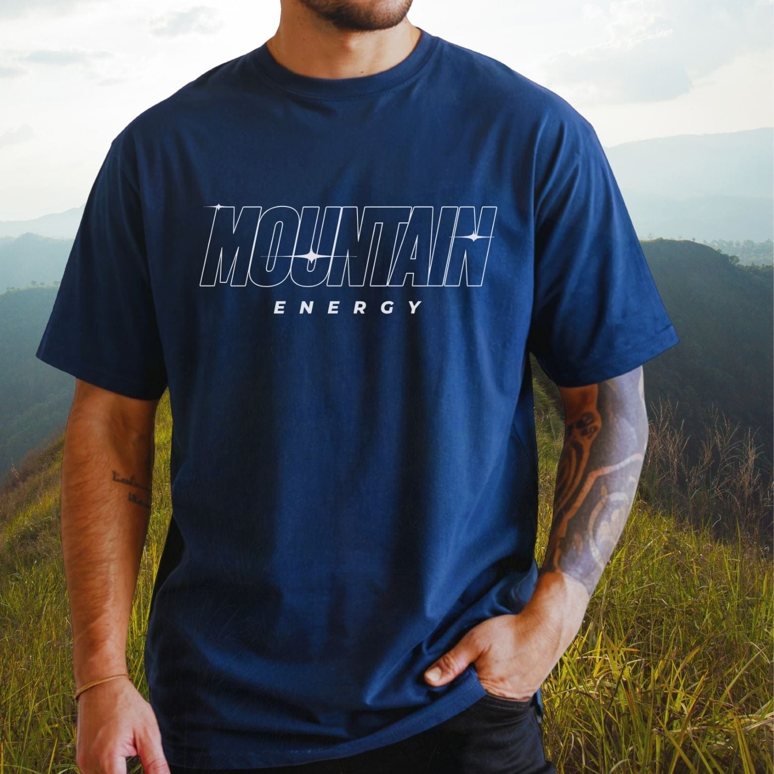 Man wearing Mountain Energy Comfort Colors 1717 Tee, showcasing its relaxed fit and soft-washed fabric, standing on a grassy hillside with mountain views.