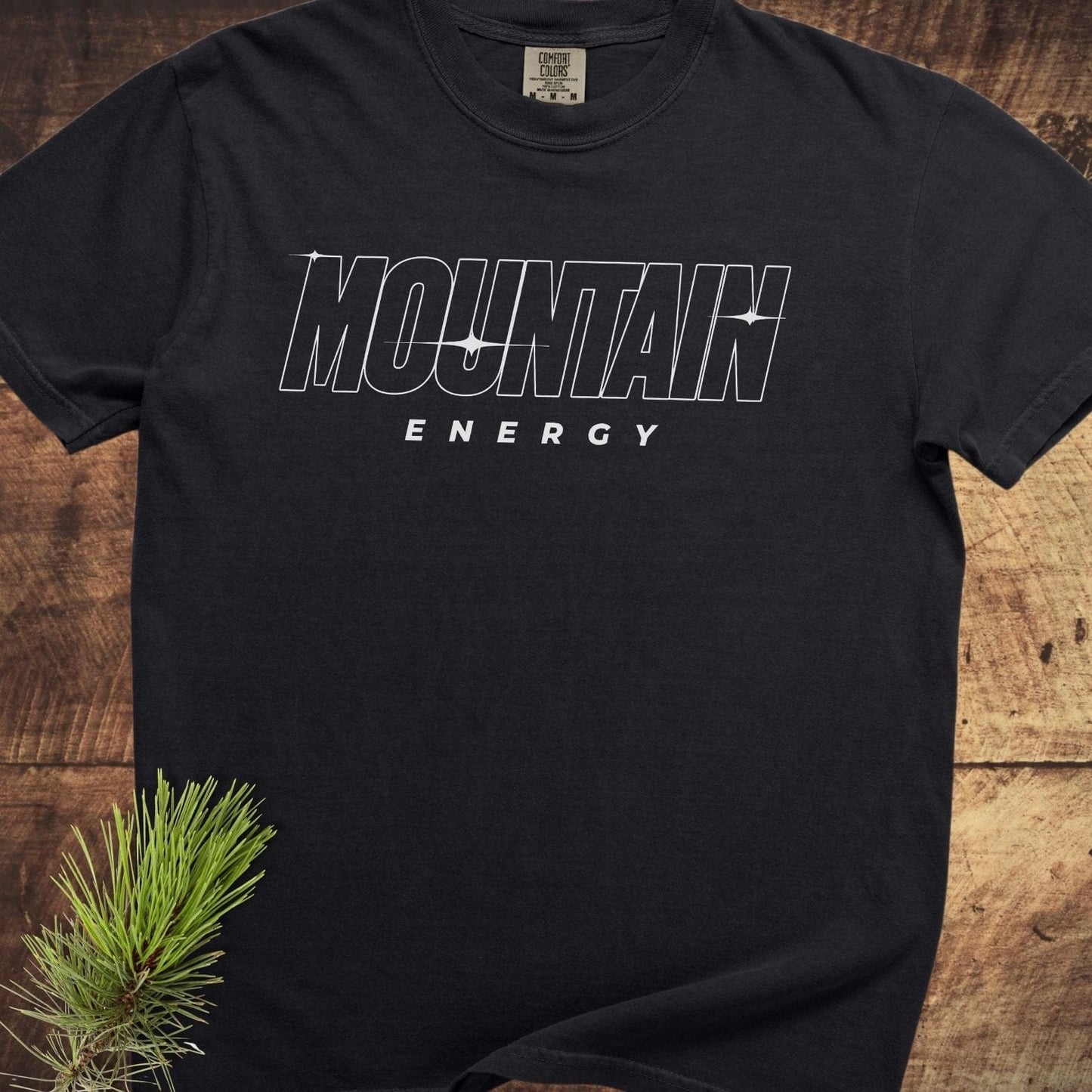 Mountain Energy Comfort Colors 1717 Tee, black, features bold white lettering, laid flat on wood. Displays MOUNTAIN ENERGY; styled with pine sprig.