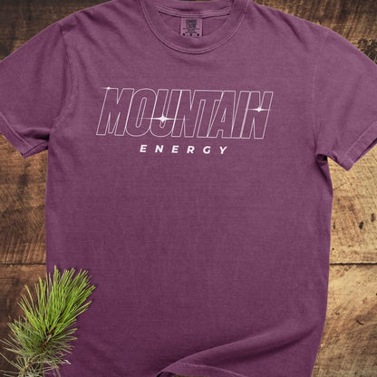 Mountain Energy Comfort Colors 1717 Tee in purple with MOUNTAIN ENERGY graphic, laid flat on wood, showcasing its soft, durable, garment-dyed fabric.