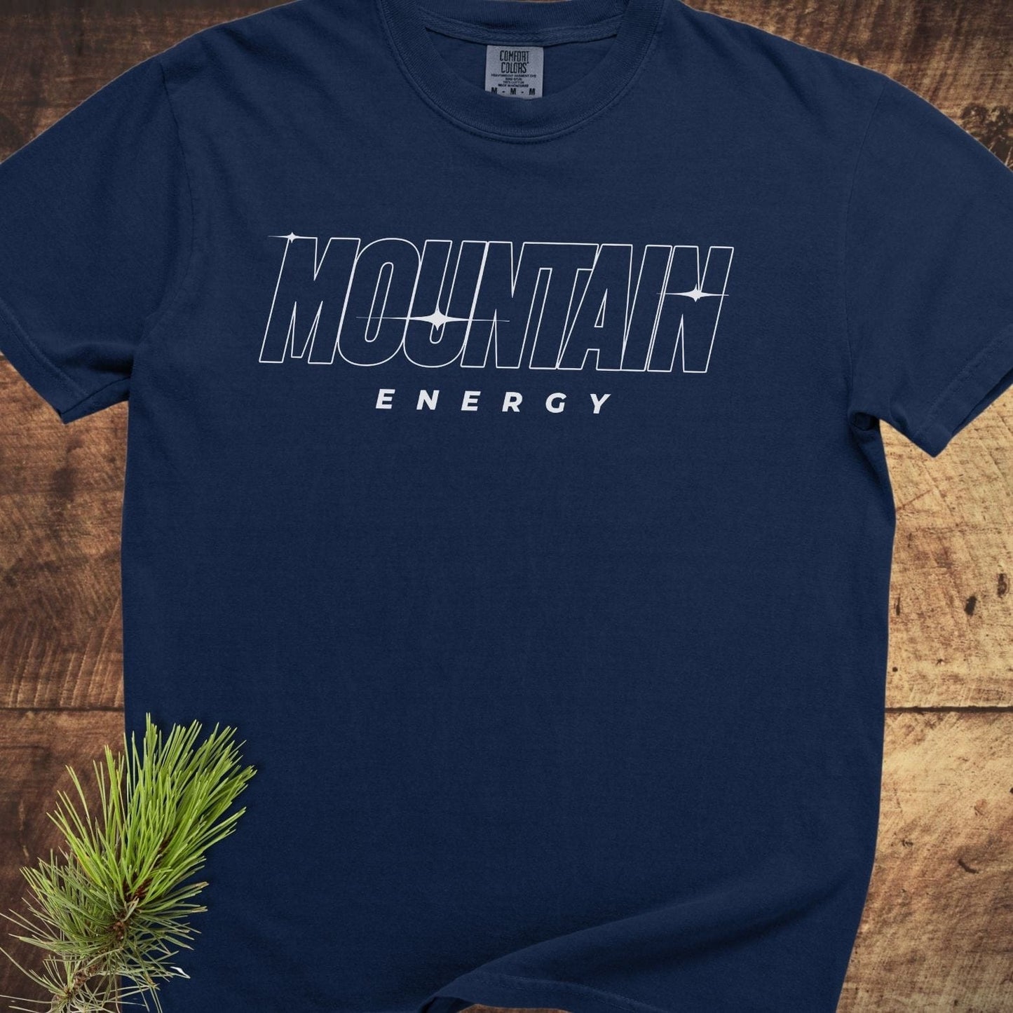 Mountain Energy Comfort Colors 1717 Tee, navy-blue, features MOUNTAIN ENERGY text, sparkle accents, and a visible neck label. Laid flat on a wooden surface.