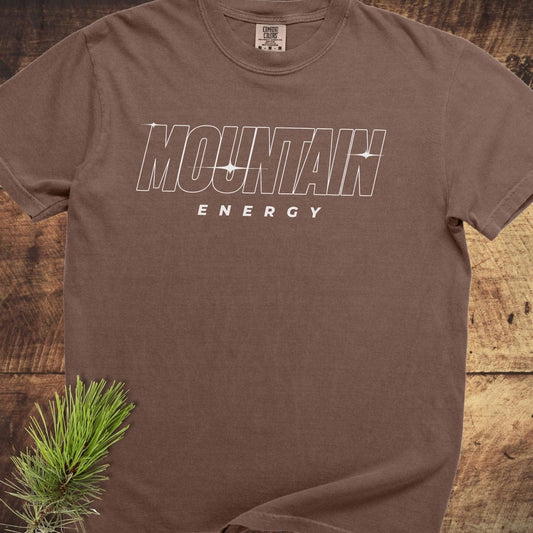 Mountain Energy Comfort Colors 1717 Tee, brown short-sleeve, crew-neck, features bold white lettering; soft-washed, 100% ring-spun cotton, laid flat on wood.