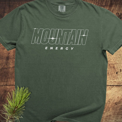 Mountain Energy Comfort Colors 1717 Tee in dark-green, featuring bold white lettering, laid flat on wood with pine needles, showcasing its relaxed fit and durable design.