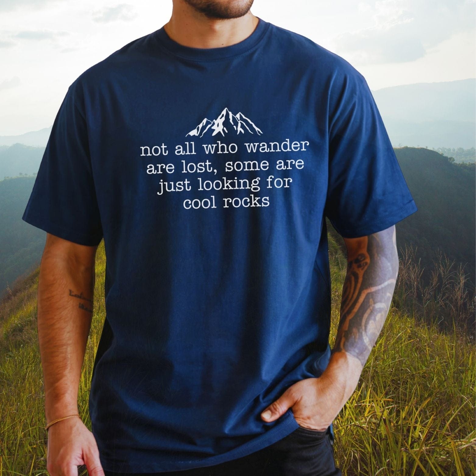 A person wears the Looking For Cool Rocks Comfort Colors 1717 Tee, featuring a mountain graphic and quote, standing on a grassy hillside.