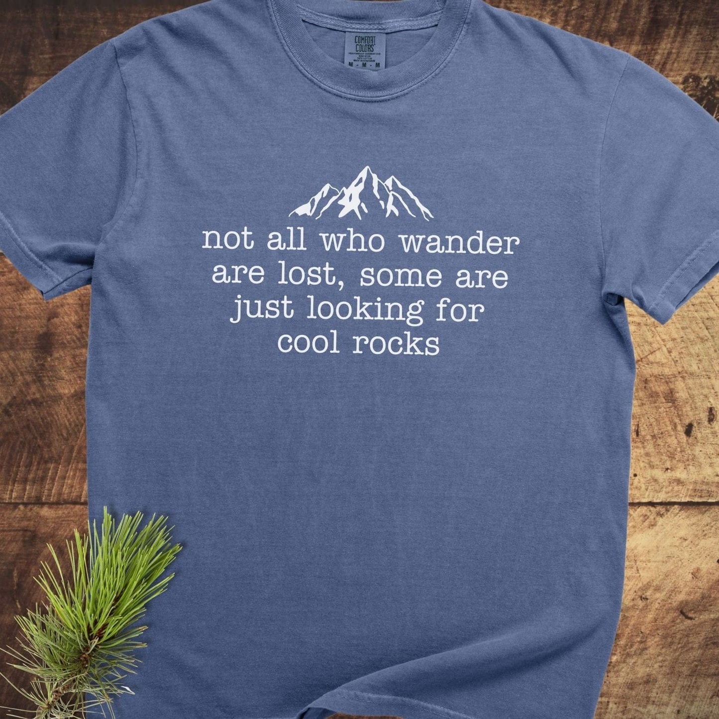 Looking For Cool Rocks Comfort Colors 1717 Tee featuring a mountain silhouette and quote, laid flat with a pine sprig, showcasing its relaxed fit and durable stitching.